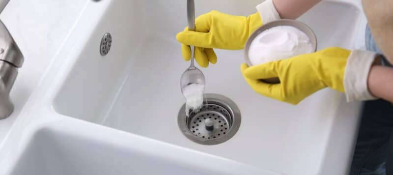 A person wearing yellow gloves pours baking soda from a spoon into a kitchen sink drain for cleaning